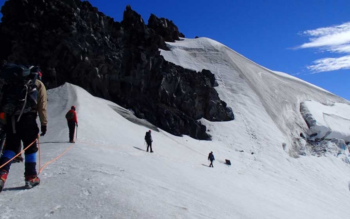 A group of people wearing safety gear are attached together by a rope, as they ascend a snowy, mountainous incline.